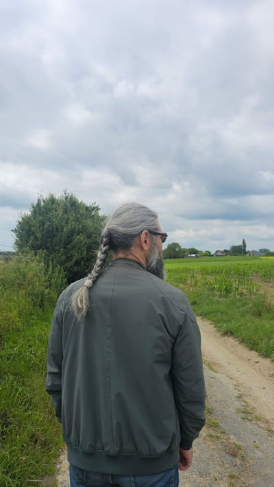 The image shows a person from behind, walking on a dirt path in a rural area. The person has long gray hair braided into a single plait and is wearing sunglasses and a dark green bomber jacket. The background features a green field, a bushy tree, and a cloudy sky. The location is identified as Silenrieux, Belgium.