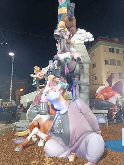 A multi-level Falla monument is centrally positioned outdoors at night in El Port De Sagunt, Spain. The monument is situated on a ground cover of brown wood chips or mulch.
In the foreground, a large, stylized male figure crouches low, wearing light purple pants, a light purple long-sleeved jacket, and a black t-shirt with "KEEP CALM AND LA CUBATA" printed in white. His face, angled upwards, exhibits light blue illumination from beneath. Behind him, a partially visible light brown and white horse figure is present, with another figure wearing a blue and white plaid cap mounted on its back, looking upwards.
Above these, a grey horse figure with pink harness details is depicted rearing, carrying a blonde female figure wearing a white dress. To the left and slightly below the grey horse, a winged cherub-like figure with blonde hair holds a scroll or book. The upper sections of the monument are supported by a grey stone-like structure adorned with white cloud formations. At the apex, a male figure dressed in green and yellow striped clothing is depicted riding a brown horse.
To the right of the main structure, another large sculptural element features a torso in an ornate red and gold vest with dark pants and large dark wings, resting on a grey base.
The background on the left reveals a crowd of people observing the monument, illuminated by a tall street lamp and other ambient lights. A large white tent structure is visible further back. On the right, a multi-story, light beige/yellow apartment building with numerous windows occupies the background, displaying faint signage including "ROOM" at ground level. The sky above is dark and clear, indicative of nighttime.