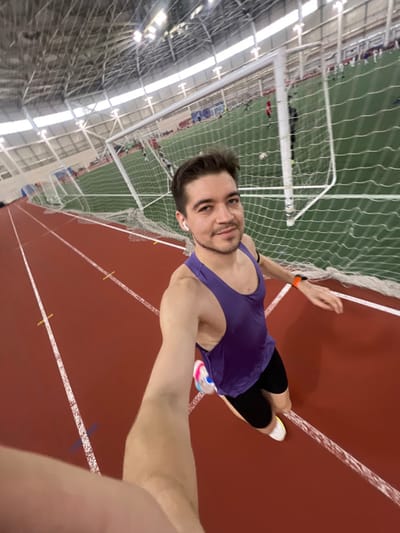 The image depicts an indoor athletic facility in Tashevka, Russia. A male individual is positioned in the foreground on a red synthetic running track marked with white lines and yellow indicators. He has short dark hair, light facial stubble, and is wearing a purple athletic tank top, black shorts, and white athletic shoes with multi-colored accents. A white wireless earbud is present in his right ear, and an orange fitness tracker is on his left wrist. He is actively taking a wide-angle selfie, extending his right arm towards the camera. In the background, beyond a white protective netting fence, lies a green artificial turf field with multiple white soccer goals. Several indistinct individuals are visible on the field, engaged in what appears to be a soccer activity. The facility's high, vaulted ceiling structure, constructed with exposed metal trusses and overhead lighting, is visible throughout the upper portion of the frame.