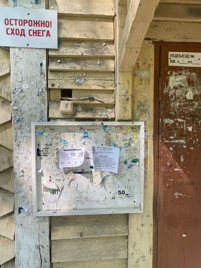 This is an outdoor shot of a weathered, cluttered bulletin board and surrounding structure, likely in Novouralsk, Russia, given the Cyrillic signage. The bulletin board, framed in white, is covered with torn notices and scraps of paper. Amongst the scattered ads, there's a notice for "Vyvoz Musora" (Garbage Removal), and an announcement mentioning the "Centralny" (Central) company and temporary disruptions in hot water quality. To the left of the board is a red and white sign warning "Ostorozhno! Skhod Snega" (Caution! Snow Coming Down). An old-fashioned electrical outlet is also visible. The wood of the structure is peeling and distressed, with numerous staples left from previous postings. The lighting suggests it is daytime and fairly bright.