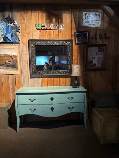 Here is a description of the image:
The image depicts a wooden wall adorned with various decorative items, presumably in an interior setting. Located in Powell, Ohio, the wall showcases a "Laundry Cleaning and Delivery Service" sign, a framed photo or document, a decorative metal piece resembling a stylized plant or branch, and a framed mirror reflecting people inside a building. Further down, the wall is decorated with a "Welcome" sign made of colorful letters, an image of blue birds and trees, and a framed photo or display board of natural exposures photography. To the right of the mirror stands a light-blue painted cabinet with drawers and black decorative drawer pulls. Above the cabinet are two wooden crates or boxes stacked on top of each other. The entire scene appears to be curated as part of an interior design or decorative arrangement.