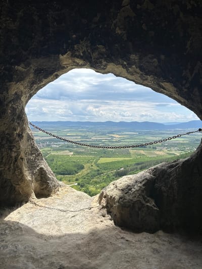 View from a cave in Han Krum, Bulgaria. The opening of the cave frames a landscape of rolling hills, green fields, and distant mountains under a partly cloudy sky. A chain hangs from the top of the cave opening, vertically bisecting the view. The interior of the cave walls are rough, textured rock formations.