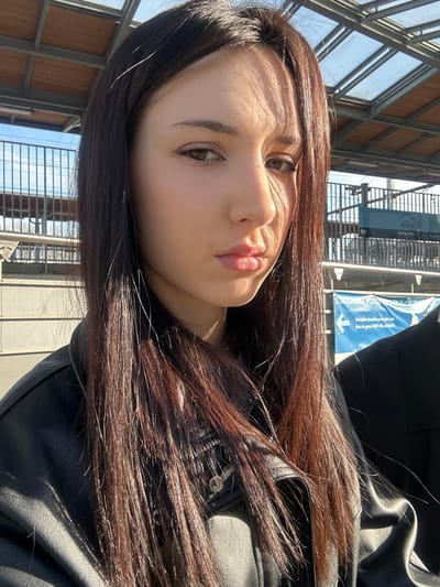 The image depicts a female subject, appearing to be in her late teens or early twenties, positioned on a train station platform in Alfortville, France. The location is confirmed by a visible blue sign reading "STATIONNEZ VOTRE" and "En gare de Alfortville." The subject has long, straight, dark brown hair and is wearing a black faux leather jacket. Direct sunlight illuminates her from the left, creating distinct shadows across her face, particularly on her right cheek and forehead. Her eyes are dark, and she is looking directly at the camera with a neutral expression, her lips slightly pursed.
In the background, overhead, is a train station canopy constructed with a metal frame and transparent or translucent panels. To the left, a metal railing and a light-colored structure are visible. To the right, a dark-clad individual is partially visible. Further back on the right, another blue sign with a bicycle icon indicates "Parking Vélo." The sky overhead is clear and blue. No specific actions or interactions are depicted beyond the subject posing for the photograph.