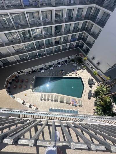 The image shows a top-down view of a hotel courtyard in Lloret de Mar, Spain. The courtyard features a rectangular swimming pool surrounded by a wooden deck. Several lounge chairs are placed around the pool, with some occupied by sunbathers. There are also tables and chairs set up on the deck, as well as potted plants and palm trees. The hotel building, with multiple stories of rooms and balconies, encloses the courtyard. The architecture is modern, with white facades and dark railings. The scene suggests a sunny day at a resort.