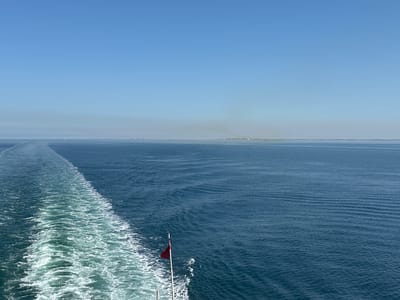 A wide-angle shot shows a vast expanse of blue sea under a clear blue sky, with a small, hazy landmass visible on the horizon. The perspective is from the stern of a moving vessel, indicated by the prominent white wake churning behind. A red and white flag is attached to a pole, possibly a Union Jack, is visible towards the bottom right of the scene. The location is likely at sea, in the vicinity of Marck, France, given the possible presence of land in the distance.