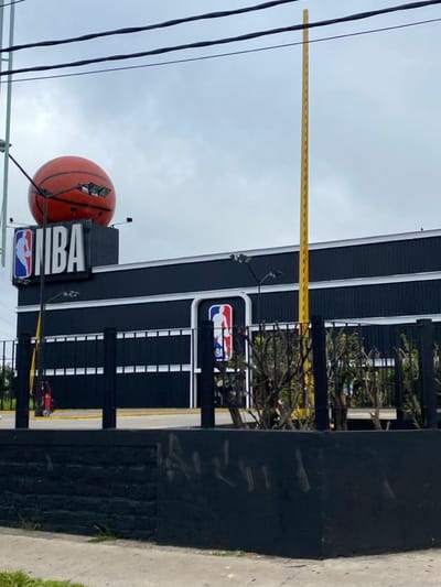 The image displays the exterior of a commercial building in Martínez, Argentina, under an overcast sky. The structure is predominantly dark-colored, featuring horizontal white banding. Mounted on the roof is a large, orange, spherical object resembling a basketball, positioned above an illuminated "NBA" sign with the standard red and blue logo. A smaller NBA logo is integrated into the building's facade at a lower level.
In the foreground, a black metal fence with vertical posts and horizontal rails runs across the scene, partially obscuring the building. Directly in front of this fence is a low, dark-painted wall exhibiting faint white markings. A concrete curb or sidewalk is visible at the very bottom of the frame. Behind the fence, a paved surface with yellow ground markings is present, along with sparse, pruned vegetation. A red, wheeled fire extinguisher is situated on the paved area to the left. A tall, slender, yellow perforated metal pole extends vertically on the right side of the image. Multiple dark utility wires traverse the upper portion of the frame. No individuals are depicted in the scene.
