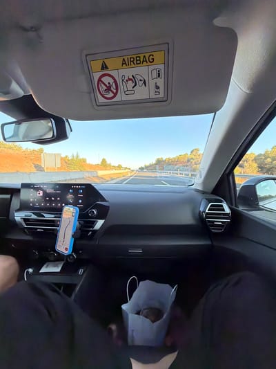 The image shows the interior of a car traveling on a highway. The dashboard features a large central infotainment screen displaying navigation, music, and vehicle information. A smartphone is mounted on the dashboard with a navigation app open. The driver's and passenger's legs are visible in the foreground, indicating at least two people are in the car. A white paper bag with some dark contents is placed on the passenger seat. The visor above the windshield has an airbag warning sticker with pictograms. The road ahead is paved, with barriers and vegetation on either side. The sky is clear and blue, suggesting daytime. The overall scene depicts a road trip experience. Based on the navigation displayed on the phone, the car is likely traveling near Requena, Spain, as indicated by the presence of road names or landmarks that are consistent with the area.
