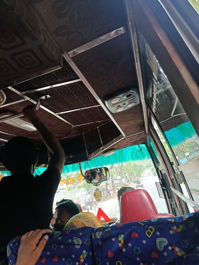 The image shows the interior of a bus in Bagerhat, Bangladesh. The ceiling of the bus is covered with brown patterned fabric and has metal beams supporting it. A person in a black shirt is seen from the back, with their right arm raised, holding onto a metal bar on the ceiling. The windshield of the bus reveals a road with trees and people on bicycles and a cart. A rear-view mirror attached to the windshield shows reflections of passengers inside the bus. In the foreground, the back of a red seat is visible, and a portion of a blue seat cover with colorful patterns is also seen. The overall scene suggests a typical commute on a bus in a Bangladeshi city.