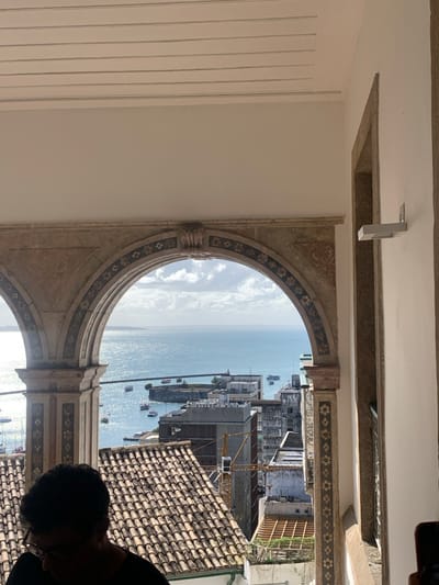 An elevated, panoramic view captures the Baía de Todos os Santos in Salvador, Brazil, framed by an ornate architectural structure. The foreground features two stone arches; one is fully visible centrally, adorned with carved details and dark floral or geometric patterns, while a second arch is partially visible on the left. Above the arches, a white ceiling with parallel linear grooves is present. To the right, a plain white wall features a horizontal, grey rectangular wall sconce.
Below the archway, multiple terracotta-tiled rooftops of city buildings descend towards the bay. The background is dominated by the expansive, shimmering blue water of the Baía de Todos os Santos under a partly cloudy sky. Numerous small boats are scattered across the bay. Along the waterfront, a circular fortified structure is visible in the mid-distance, surrounded by various urban buildings, including a prominent yellow construction crane.
In the lower-left foreground, the upper portion of an individual's head and shoulders is visible. The person has dark hair, wears glasses, and a dark shirt, appearing to look downwards out of frame.