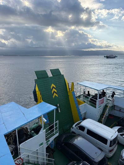 The image shows a view from a ferry in the ocean near Melaya, Indonesia. The sky is partly cloudy with sunbeams breaking through, creating a dramatic lighting effect over the water. In the distance, a coastline with hills or mountains is visible. A smaller ferry boat is seen on the water.
The foreground of the image is dominated by the deck of the ferry the photographer is on. There are cars parked on the deck, including a white van and at least two other cars. Ropes and winches are visible on the deck, suggesting the ferry is used for transporting vehicles. A green and yellow ramp or gate is partially raised, indicating it may be preparing to dock or depart. Several people are visible on the ferry's deck, some standing and observing the surroundings. The ferry has blue awnings providing shade. The overall scene depicts a ferry journey in a coastal environment.
