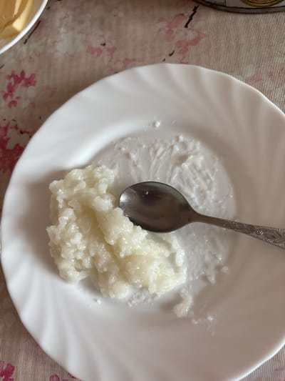 A white plate containing a pile of cooked rice and a metal spoon sits on a table in Orel, Russia. A small amount of rice is smeared on the plate's surface. The table is covered with a patterned tablecloth. Part of a dish with a yellow substance is visible in the lower-left corner of the image.