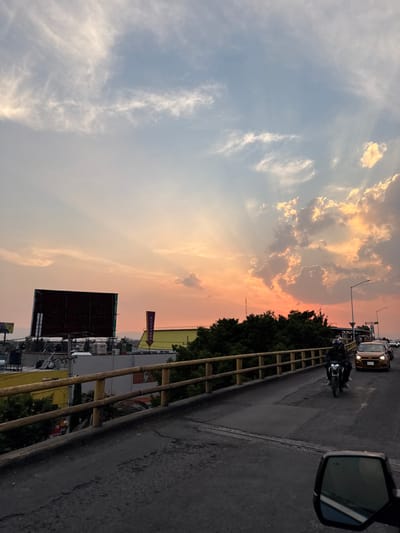 The image depicts an elevated view of an urban environment in Nezahualcóyotl, Mexico, at sunset. The sky displays a gradient from light blue with wispy clouds at the top to vibrant orange and pink hues near the horizon, with prominent crepuscular rays and illuminated cumulus clouds.
In the foreground, the dark gray asphalt surface of an elevated road or overpass is visible, with a vehicle's side mirror partially obscuring the lower right corner, indicating the photograph was taken from a vehicle. A yellow-painted concrete railing runs along the right side of the elevated structure.
On the road, a dark-colored motorcycle, ridden by a person wearing a helmet, is traveling away from the viewer. Behind the motorcycle, a line of cars is visible, including a light brown/orange sedan, also moving in the same direction.
Below and to the left of the elevated road, various buildings with flat roofs and some yellow facades are visible, partially obscured by green trees and foliage. Prominent features include two large, unlit rectangular billboards on rooftops and a tall, slender purple sign shaped like a downward-pointing arrow, bearing white text that appears to be "FRESKO" or "FRESKI". Distant urban structures and hazy hills are faintly discernible on the horizon beneath the sunset glow. Streetlights are visible but unlit.