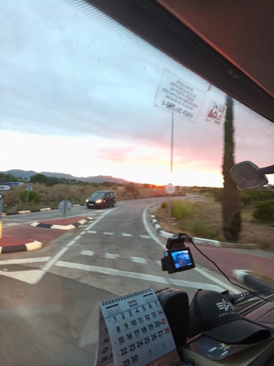The image offers a first-person perspective from inside a moving vehicle, looking out through the windshield at a sunset scene. The sky is illuminated with soft orange, pink, and pale blue colors as the sun dips below the horizon, casting a warm glow over distant hills or mountains.
Outside the vehicle, a winding road, possibly leading to a roundabout or intersection, is visible. A dark-colored car is seen ahead, navigating the road. Roadside vegetation, including dry grasses and a distinctive tall, slender tree resembling a cypress, lines the route. Road markings, a small reflective element on the road, and road signs are also present.
Inside the vehicle, a dashcam is mounted on the windshield, with its screen displaying an active recording. Below it, on the dashboard, rests a desk calendar for "Junio 2026," clearly showing all days of the month, with some dates like 14, 21, and 28-30 being particularly legible. Next to the calendar, a black baseball cap with the "ALPHA INDUSTRIES" logo is placed.
Notable details include a sticker on the windshield displaying "SPEED LIMITER," "LIMITADOR DE VELOCIDAD," and "V-SET=90 KMH," along with another smaller red and white triangular sign showing "40." The scene evokes a peaceful drive during dusk in a rural or semi-rural area, consistent with the location context of Nàquera / Náquera, Spain.