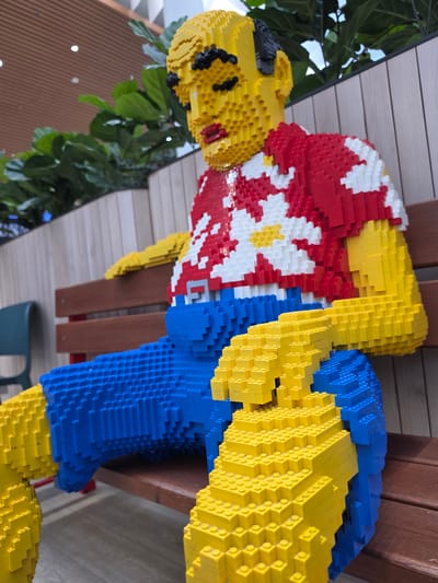A life-sized statue of a person constructed entirely from yellow, red, and blue Lego bricks sits on a wooden bench. The figure wears a red shirt with white floral patterns and blue shorts. Its face is yellow with black eyes and red lips. The statue's legs are crossed, with one arm resting on its lap and the other extended outwards. The background features lush green foliage and a wooden wall. This statue is located in the city of Mladá Boleslav, Czechia.
