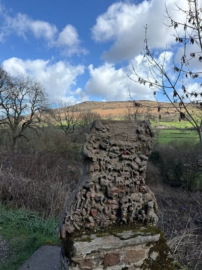 The image displays a weathered, intricately carved stone sculpture, possibly concrete, resting on a moss-covered stone and brick plinth in a rural setting within the High Peak area of the United Kingdom. The sculpture, which appears columnar, is densely decorated with numerous small, low-relief figures and patterns, exhibiting a dominant grey-brown coloration with sparse, faded red accents. To its immediate left is a narrow, light-grey path bordered by short green grass. The midground surrounding the sculpture is characterized by dense, dry, tangled brown shrubbery and several large, bare deciduous trees with dark, gnarled branches. In the background, a rolling landscape unfolds under a bright blue sky dotted with white cumulus clouds. A prominent hill or moorland covered in light brown, dry vegetation occupies the upper background, while sections of vibrant green fields and faint distant structures are visible below it. A bare tree branch with dark, dried seed pods extends into the frame from the upper right. No persons are present.