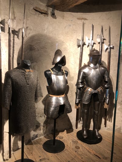 The image shows a display of medieval armor and weapons inside what appears to be a museum or historical site, possibly in the city of Veytaux, Switzerland. There are three sets of armor arranged from top to bottom. The top set is a complete suit of plate armor on a stand, consisting of a helmet, breastplate, gauntlets, and leg protection. Below it is a partial set of plate armor, including a helmet and breastplate on a stand. The bottom set is a chainmail shirt on a mannequin. Various polearms are displayed against the back wall behind the armor, including halberds with distinctive axe-like blades and spear tips. The backdrop is a rough, uneven stone wall. The display is positioned on a wooden floor. The lighting appears to be artificial and casts shadows.