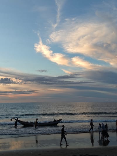 At sunset in Peuneulet Baroh, Indonesia, silhouetted figures navigate the shallows of a beach. Two individuals are pushing a long, narrow fishing boat into the gentle waves, one holding a paddle or oar. Further down the beach, a group of people, including children, are walking along the water's edge. The sky is painted with a gradient of blues and oranges, with wispy, illuminated clouds suggesting the fading light. The wet sand reflects the muted colors of the sky and the figures. The ocean is calm with small waves lapping at the shore.