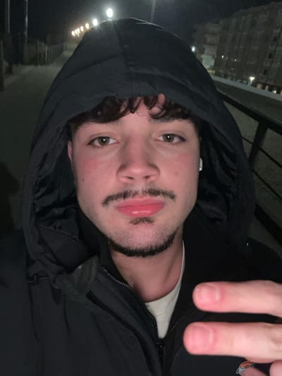 A person with a hooded jacket poses for a photo at night. The individual has short, curly dark hair, brown eyes, and stubble. Two fingers are held up in the top right corner. The background shows blurry lights. The jacket is a dark color, possibly black. The person's face is slightly flushed. An earbud is visible in the left ear.