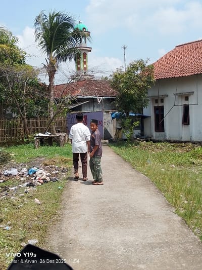 Two people walk along a concrete path lined with vegetation. One wears a white shirt and brown pants, the other a patterned shirt and pants, both with head coverings. A palm tree and a building with a green dome rise above the trees. A house with a red tile roof is on the right. Rubbish litters the left side of the path. A phone camera overlay is visible in the lower left corner of the frame.