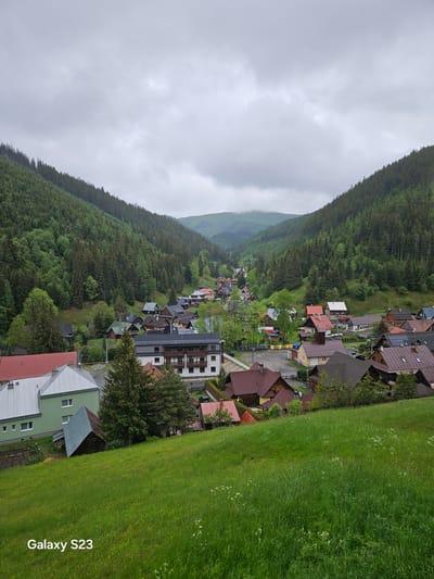 The image captures an aerial view of Vyšná Boca, Slovakia, on an overcast day. The village is nestled in a valley, flanked by densely forested hills. A variety of buildings, predominantly houses with red and brown roofs, are scattered along the valley floor. A road winds through the village. Patches of bright green grass contrast with the darker shades of the forest. The sky is heavily clouded, casting a soft light over the scene. The bottom right corner of the image displays the text "Galaxy S23".