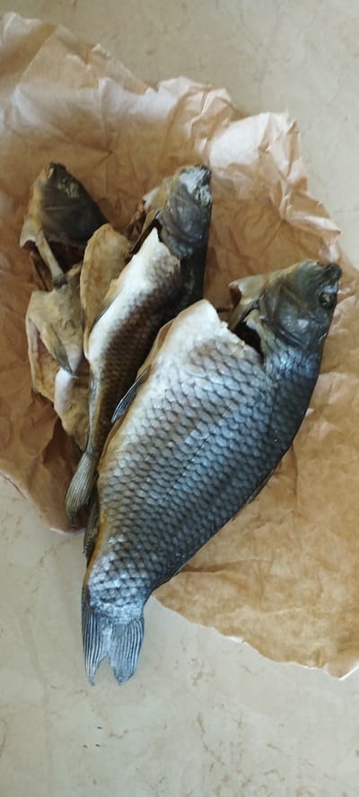 A close-up, overhead shot shows several dried fish laid out on crumpled brown paper, which rests on a light-colored, marbled surface. The fish are whole, but appear to be split open, with their interiors exposed. Their scales are visible, and they have a greyish-blue hue. One fish is positioned prominently in the foreground, with its tail extending towards the bottom left of the frame. The lighting is even, and there are no people or animals visible in the scene. The specific location of Skidzieĺ, Belarus cannot be determined from the visual information provided.