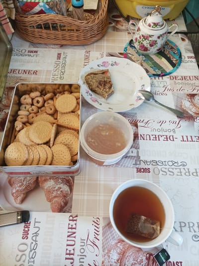 This media file depicts a top-down view of a table set for breakfast or a tea break, likely in a home setting. The table is covered with a tablecloth featuring a repeating pattern of French breakfast terms like "SUCRE," "CROISSANT," "JEUNER," and "Bonjour," interspersed with illustrations of pastries.
Prominently displayed are a tin filled with various types of cookies and a white ceramic teapot adorned with floral patterns. Next to the teapot, a small, colorful quilted coaster is visible. A partially eaten piece of what appears to be a pastry or crêpe sits on a white plate, accompanied by a spoon. A clear plastic container holds a golden-colored substance, likely honey or jam. In the foreground, a white mug contains dark tea with a tea bag, and the "Greenfield" brand of tea is visible on the tag. There are also two croissants dusted with powdered sugar on a plate, and a metal grater to the left. In the background, a woven basket holds more food items, possibly cookies or snacks, and a yellow appliance.
The overall scene suggests a peaceful and leisurely moment. The lighting and the presence of breakfast items imply it is morning. There are no people visible. The text on the tablecloth, teapot, and tea tag indicates a likely European setting, with the "Прыбыткаўскі сельскі Савет, Belarus" location context further narrowing it down. The visual arrangement of food and drink evokes a sense of comfort and indulgence.