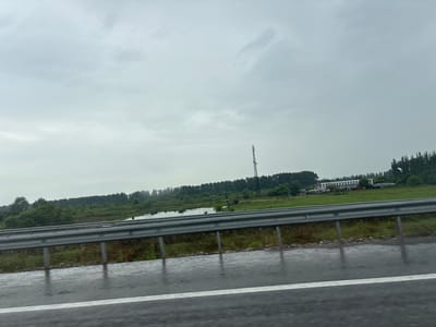 The image depicts a roadside view on a cloudy day. A guardrail runs horizontally across the foreground, separating the wet asphalt road from a grassy embankment and a body of water. Beyond the embankment, a line of trees forms the horizon, with a tall tower structure visible in the distance. Agricultural buildings are also present among the trees. The scene appears to be on the outskirts of a city, possibly in an industrial or rural area. The weather is overcast, and the road surface indicates recent rain. There are no people or vehicles visible in the frame. The location is identified as ქალაქი ლანჩხუთი, Georgia.
