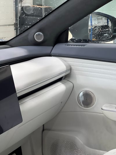 The image shows the interior of a modern car, focusing on the door panel and dashboard. A silver speaker grille is visible on the door panel, and another smaller speaker grille is mounted on the A-pillar near the windshield. The interior features white and dark gray upholstery, with textured panels and a metallic trim accent on the dashboard. A side mirror is reflected in the windshield, showing a blurred image of the exterior. The scene is likely in a workshop or showroom, as suggested by the presence of stacked tires visible through the windshield and the overall clean and staged appearance. The location of Kabanikha, Russia, is not discernible from the image itself, nor are there any people or specific actions depicted.