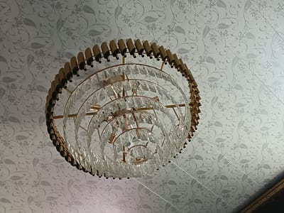 The image shows a chandelier hanging from a patterned ceiling. The chandelier is made of brass and has multiple tiers of clear crystals. The ceiling has a light grey floral pattern. The image is taken from a low angle, looking upwards. There are no people or actions depicted in the image. The location is specified as Uchkun, Kyrgyzstan, but there are no visual cues in the image to confirm this location.