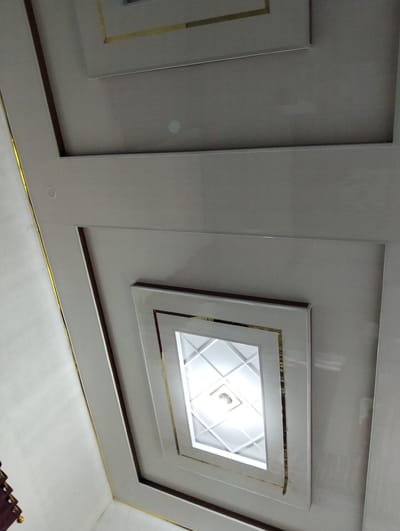 This is an eye-level, close-up shot of a modern ceiling, taken indoors in Aluedua, Indonesia. The ceiling features a stepped, recessed design with a central illuminated panel. The recessed areas are framed with thin, gold-colored trim, adding a touch of elegance. The main surface of the ceiling appears to be a smooth, light-colored material, possibly PVC or a similar synthetic finish, with a subtle sheen. The central panel is a bright white, with a geometric, diamond-shaped pattern created by intersecting lines. A light fixture is visible within this central panel, emitting a bright, diffused light. The overall impression is one of contemporary interior design, likely in a residential or commercial space.