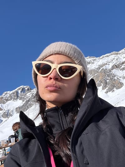 A female subject is depicted in a close-up photograph, set against a backdrop of snow-covered mountains and a clear blue sky in Klösterle, Austria. She wears a light beige knitted beanie and cream-colored cat-eye sunglasses with dark lenses, featuring a "Ray-Ban" logo on the left lens. Her dark hair is styled in two braids, visible over her shoulders. She is dressed in a black zippered outer jacket, a black zippered mid-layer, and a bright pink inner garment. Her face is illuminated by sunlight, and her lips are pursed. To the left and slightly behind the main subject, a second individual is partially visible, seated at a wooden picnic table with several white cups. This background figure wears a dark jacket and a light-colored face covering. The scene indicates a bright, sunny winter day in an alpine environment.