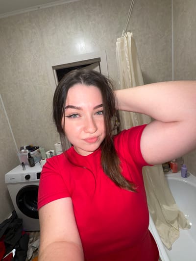 This image, taken in Kitsjanovo, Russia, captures a young woman posing for a selfie in a domestic bathroom. The photo is oriented 90 degrees clockwise from a standard portrait view.

The main subject is a woman with long, dark brown hair, striking blue-green eyes, and full lips, who is looking directly at the camera with a calm and slightly smiling expression. She is wearing a bright red short-sleeved top. Her bent legs are visible in the upper right quadrant of the image, suggesting she might be lying in a bathtub or on the bathroom floor.

The bathroom features light-colored, subtly patterned tiled walls. To the right, a white bathtub contains various plastic bottles of toiletries on its edge, next to a beige shower curtain that is partially drawn. Below the bathtub, a white front-loading washing machine, branded "Gorenje", is visible. On top of the washing machine, several rolls of toilet paper are stacked, alongside a small container holding miscellaneous items. A pile of dark clothes is situated next to the appliance. A rectangular vent or window frame is visible on the wall to the left of the woman's head. The lighting appears to be artificial, typical of an indoor bathroom setting.