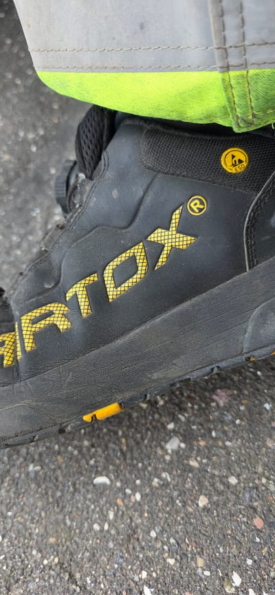 The image is a close-up, low-angle shot of a black work boot with yellow branding. The boot is dirty and shows signs of wear. The branding consists of a large, stylized yellow word that appears to be "ARTOX," with a smaller registered trademark symbol to its right. Above the "ARTOX" logo, there is a yellow circular emblem with a symbol resembling a lightning bolt inside a circle, and the letters "ESD." The boot is resting on a rough asphalt surface. In the background, a portion of a high-visibility garment, likely a jacket or vest, is visible. It has a gray upper section with reflective stitching and a bright neon green lower section. The image does not provide any specific geographical landmarks or context to determine its location within Hjørring, Denmark.