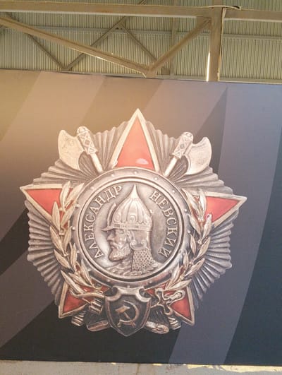 The image shows a close-up of a large medal featuring a profile of a man in a helmet, surrounded by laurel branches and crossed axes. The medal is predominantly silver with red accents. Cyrillic lettering is visible around the central portrait. Below the medal is a shield with a hammer and sickle emblem. The medal is displayed against a dark background with a geometric pattern of light and dark gray stripes. Above the medal, a corrugated metal ceiling and industrial metal beams are visible, suggesting an indoor setting. The overall impression is of a museum exhibit or public display. The medal is likely the Order of Alexander Nevsky, a Soviet and Russian award. The image itself does not contain any visual information to pinpoint its location in Pesochny, Russia.