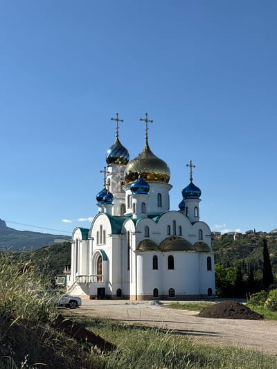 A white orthodox church with multiple blue and gold domes topped with crosses stands against a clear blue sky. The church is situated on a hillside with lush green vegetation and trees. In the foreground, tall dry grass and a dirt pile are visible. A white car is parked near the church. The scene appears to be in a rural or semi-rural area, possibly a village, given the presence of houses on the distant hillside. The image is taken in daylight, with the sun casting shadows. The overall impression is one of tranquility and religious architecture against a natural backdrop.