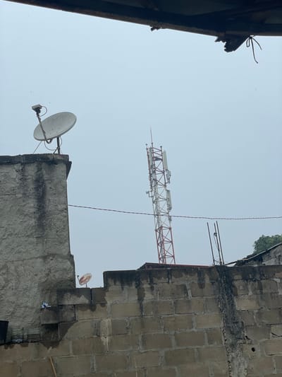 This image presents an upward-looking view of an urban landscape in Luanda, Angola, dominated by an overcast sky. The central subject is a large, red and white lattice tower crane, stretching horizontally across the middle of the frame. Several rectangular components are mounted along its length, possibly part of its communication or operational apparatus. The crane appears somewhat distant, its details softened by the grey, diffused light.
In the foreground, portions of concrete buildings frame the scene. On the left, a dark, angled edge of a structure with visible damage and a bent wire points into the sky. On the right, a multi-storied building constructed from large, grey concrete blocks shows signs of weathering and dark stains from moisture. A small cluster of green foliage is visible atop this building section.
Two satellite dishes are mounted on the lower part of the right-hand building. A prominent, round white satellite dish is fixed to the structure, pointing towards the left, while a smaller, reddish dish is partially visible further to the right. A thin, dark wire or cable hangs vertically through the center-right portion of the image. The sky is a uniform, bright light grey, suggesting a cloudy day without direct sunlight. There are no people visible, nor any discernible text. The scene conveys a sense of ongoing urban development or activity, characteristic of a bustling city.