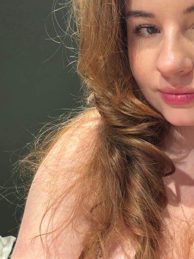 The image shows a close-up of a woman's face and shoulder, with her auburn hair framing the left side of the frame. She is looking to the right, and her makeup is visible. The background is a dark, solid color, likely a wall. The lighting appears to be artificial and casts shadows on her skin. There are no identifiable landmarks or elements that would suggest a specific location within Fredrikstad, Norway.