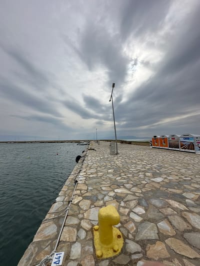 This image depicts a pier in Limenas Avdiron, Greece, under an overcast sky. The foreground features a yellow, T-shaped mooring bollard bolted to a light-colored, irregularly paved stone surface. To the left, dark, rippled water contains several orange and yellow buoys, and multiple small boats are moored further along the pier. Black cylindrical fenders hang along the pier's edge, and a rope with a white label marked "Δ41" is visible near the foreground bollard. A tall, slender streetlight with an integrated fixture stands in the mid-ground, leaning slightly to the left, with additional similar lights extending into the distance along the pier. On the right side of the pier, a row of four large, multi-compartment recycling bins, each with distinct colors and graphics, is visible. Beyond the bins, a light-colored bench and a low wall border a grassy area. Distant land or hills are visible on the horizon beneath the grey, cloud-laden sky, which shows a brighter patch where the sun partially penetrates. No individuals or specific actions are depicted.