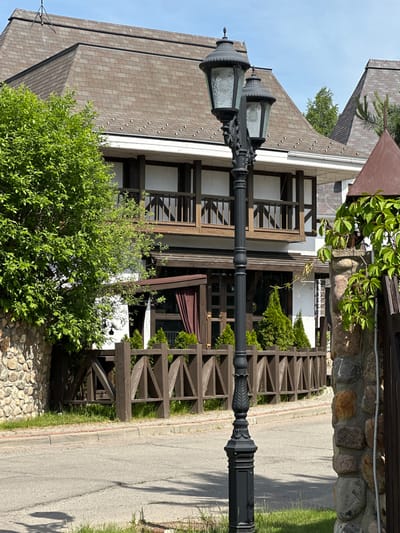 This image captures a street scene in NovoGlagolevo, Russia, under bright daylight. A prominent two-story building with a multi-gabled brown shingle roof dominates the background. The roof is equipped with numerous small, white snow/ice guards. The building's facade is white, accentuated by dark brown wooden beams, window frames, and balcony railings on the upper floor. On the ground floor, a large window is visible, covered by a dark brown curtain, and flanked by several small, conical evergreen shrubs. A dark brown wooden fence with a distinctive triangular pattern encloses this ground-level area.
In the mid-ground, a tall, black, ornate cast-iron street lamp stands centrally. It features a decorative base and shaft, topped with two glass lanterns with textured or frosted glass. To the left of the scene, a large, vibrant green deciduous tree partially obscures the building's lower section and a segment of a rough-hewn stone wall constructed from various sizes and colors of natural stones. To the right, a similar stone pillar, partially covered in green ivy, stands beside a dark brown wooden gate structure. The foreground features a gray asphalt road with visible cracks and seams, bordered by a reddish curb and patches of green grass. Shadows are cast by the street lamp and other elements, indicating a strong light source from the upper right.