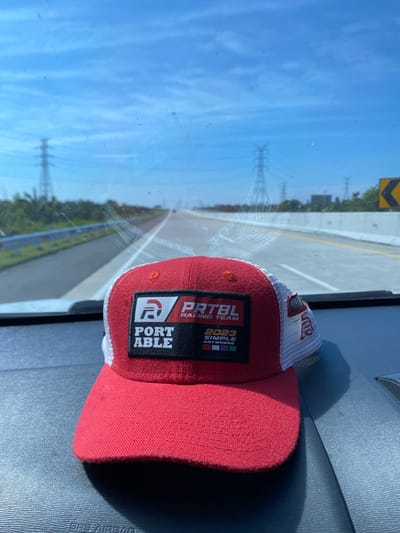 A red and white trucker hat with a black patch featuring "PORTABLE RACING TEAM" and "2023 SIMPLE ANYWHERE" is resting on a gray dashboard inside a vehicle. The hat's front is red, while the back features white mesh. The background visible through the windshield shows a highway under a clear blue sky with some wispy clouds. Power lines and transmission towers are visible in the distance along the highway, suggesting a location outside a dense urban center, possibly in the outskirts of Stabat, Indonesia, given the presence of extensive power infrastructure typical of developing industrial areas or transportation corridors in such regions. The road appears to be a well-maintained highway with concrete barriers and yellow lane markers.