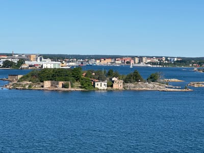 The image depicts a scenic view of the city of Karlskrona, Sweden, from the water. In the foreground, a small, rocky island is visible, dotted with a few weathered stone structures and some vegetation. Further in the background, the cityscape of Karlskrona unfolds along the coastline, characterized by a mix of modern and older buildings, including residential and commercial structures, and a harbor with various boats and ships. The water is a deep blue, reflecting the clear, bright sky. Trees and foliage are present along the shoreline and on the island. The scene appears tranquil and offers a glimpse of urban life integrated with the natural coastal environment of this historic Swedish city.