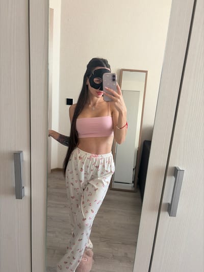 A female individual is depicted taking a mirror selfie in an indoor domestic setting in Krasnaya Zarya, Russia. She wears a black eye mask, a light pink spaghetti-strap crop top exposing her midriff, white pajama bottoms with a pink floral pattern and a pink bow, and light pink fuzzy slippers. Her long dark hair extends over her left shoulder, and a tattoo is visible on her right arm. She wears a silver pendant necklace and a red string bracelet on her left wrist. She holds a light purple smartphone with a clear, embellished case in her right hand, pointed at her reflection. The environment includes a light-colored wall, light wood-grain wardrobe doors with grey handles framing the primary mirror, and light wood flooring. A second, freestanding mirror with a wooden frame is visible in the background reflection, reflecting a dark furniture item.