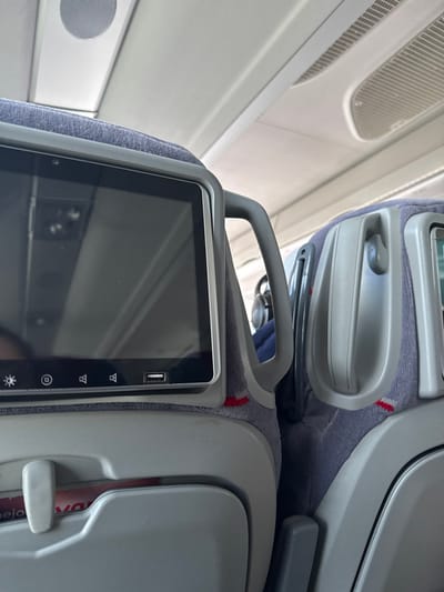 The image provides an interior view of an aircraft cabin, focusing on the back of a passenger seat and its in-flight entertainment (IFE) system. In the foreground, a modern IFE screen is prominent, mounted on the seatback which is upholstered in a grey fabric with faint red stitching. The screen features a dark display and clearly shows a USB port at the top right. Along the right edge of the screen, several touch-sensitive icons are visible, including one resembling a sun for brightness control. An integrated grey handle is positioned above the screen.

To the right of the main screen, part of a folded tray table or a storage compartment is visible, with a very small portion of red text, possibly "OVO" or "OV," just discernible on an item within it. Above the foreground seat, another seat with a similar IFE screen setup is partially seen, suggesting a row of seating. The upper left of the image reveals the aircraft's white overhead cabin panels, complete with ventilation grilles and a circular speaker.

The overall lighting is bright and even, characteristic of an aircraft cabin, possibly during daytime. The image is captured from a slightly tilted perspective. No people are visible. The setting is clearly an enclosed aircraft environment, and there are no specific visual cues within the image to the provided location context of Guadarrama, Spain.