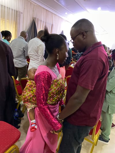 A medium, indoor shot shows a gathering of people in a decorated space. The lighting is bright and overhead. In the foreground, a woman with dark hair pulled back is seen in profile wearing a bright pink outfit with a patterned green and red overlay. She wears silver earrings and has fair skin. A man with glasses, a bald head, and a burgundy shirt is bending over her. Several people sit behind them, including a man in a white shirt and another man in a light green traditional outfit. The environment suggests an event, with draped fabrics, gold chairs, and people dressed up. The city of Miango, Nigeria, is mentioned as the location.