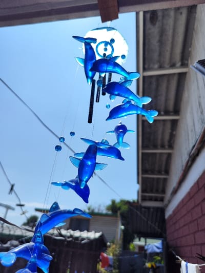 A dolphin wind chime hangs outside. The bright blue dolphins are suspended by clear strings with small blue beads. The top is a circular piece with a pattern cut out, and there are also several black tubes hanging down. The background includes a blue sky, the roof of a house, a brick wall, and green trees. The scene is brightly lit.