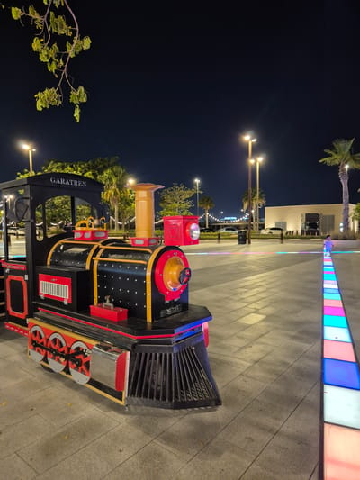 A nighttime exterior scene in Fujairah, United Arab Emirates, captures a toy train attraction positioned in a wide, paved public area. The train is black with red and yellow accents and features the name "GARATREN" emblazoned on its roof. In the background, palm trees and streetlights illuminate the area, with a building and distant lights visible further back. A young child is walking on a pathway lined with illuminated, multi-colored square tiles. The sky is dark, indicating it is nighttime.
