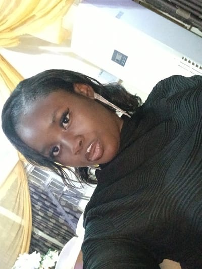 A young adult female, with dark skin and dark hair styled with braids and wavy extensions, is depicted in a close-up, tilted portrait. She wears a black, textured, long-sleeved top and a prominent, dangling, silver-toned earring on her visible right ear. Her face features winged eyeliner and glossy, parted lips, with her tongue slightly visible, as she gazes directly at the camera. The background indicates an indoor setting, likely an event venue, featuring yellow draped sheer fabric on the left, partially visible white floral arrangements in the lower left, and a dark patterned fabric behind the subject's left shoulder. To the upper right, a white wall or panel is visible, upon which a dark rectangular control panel with a digital display is mounted, adjacent to horizontal vents. The image does not contain identifiable markers to confirm the location as Isasi, Nigeria.