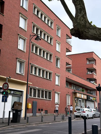The image captures a street scene in Maisons-Alfort, France, featuring a multi-story brick building with numerous windows and balconies. A large tree branch partially obscures the upper portion of the building on the right side. In the foreground, a sidewalk runs alongside the building, with bollards lining its edge. A pedestrian crossing is visible in the lower part of the frame.
On the sidewalk, several individuals are present. Two men and two women are walking away from the viewer. A black trash bin, labeled "Paris," is positioned near the sidewalk. A street light with a pinkish globe is also visible. To the left, a traffic signal shows a green walking man icon and an upward-pointing blue arrow sign. Below this, a circular red and white sign displays "7,3m".
Further down the sidewalk, there are shops with closed storefronts. One shop has a sign that reads "Ranch Chicken Fried Chicken." Two cars, a white station wagon and a silver sedan, are parked in front of these establishments. A yellow mailbox is mounted on the brick wall of the building. A gray door is also visible on the building's facade. The overall environment suggests a moderately urban setting.