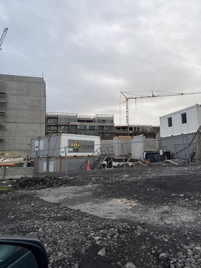 This image depicts an ongoing construction site in Reykjavik, Iceland, likely a new building or complex. Dominating the scene are several large concrete structures, indicative of new residential or commercial buildings under construction. Two prominent construction cranes are visible, one a dark, latticed tower crane on the left and a bright orange lattice boom crane in the distance on the right, both essential for lifting and moving heavy materials on such a site.
In the foreground, several modular temporary structures, commonly used as site offices or storage units on construction projects, are parked. One of these units features a white side panel with a logo that appears to be "GUNNAR BJARNARSON EHF", suggesting the name of a company involved in the construction or development. The ground surrounding the site is covered with dark, rough gravel and rocky material, typical of undeveloped or industrial areas in Reykjavik. A temporary metal fence encloses parts of the construction zone, a standard safety measure.
The sky is overcast with grey clouds, creating a diffused, somewhat dull lighting condition that is characteristic of Icelandic weather. The overall impression is one of active development and infrastructure expansion within the city. There are no visible people in the image.