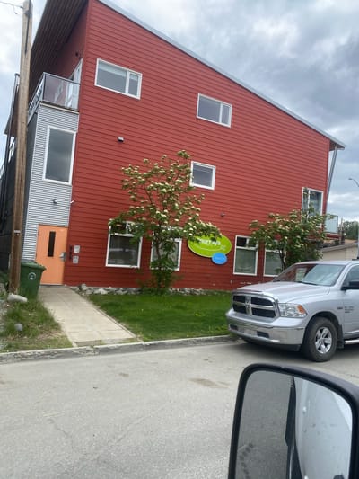 A silver Dodge Ram 1500 pickup truck is parked on a paved road in front of a two-story building with red siding and a sloped metal roof. The building has several windows and an orange door on the ground floor. A green trash bin is next to the door. There is a sidewalk and a grassy area between the building and the road. A green oval sign with white lettering is mounted on the building. The sky is overcast. The location is the Murray Dental clinic at 4969 4th Avenue in Whitehorse, Canada.